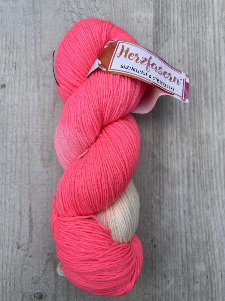 Ein Strang handgefärbter Wolle in kräftigem Neon-Pink mit sanften, hellen Übergängen ins Cremefarbene. Die Banderole trägt das Herzfasern-Logo mit der Aufschrift „Garnkunst & Strickliebe“. Der Strang liegt auf hellem Holzuntergrund.